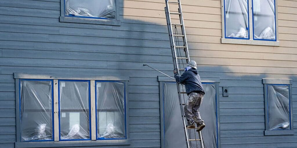 Exterior painter on ladder inspecting siding to show how winter damage shows up on exterior paint surfaces through peeling and wear