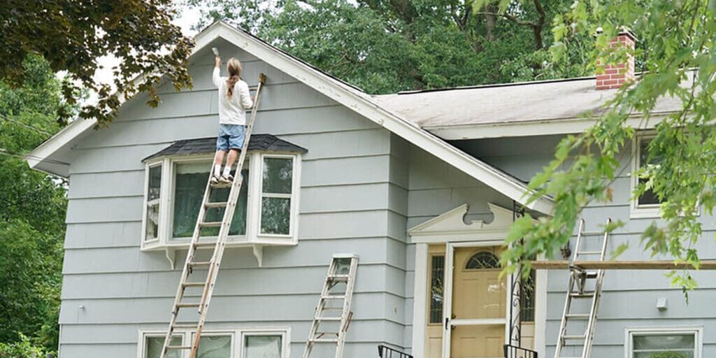 Homeowner on ladder painting house exterior, illustrating when should you start planning exterior painting in Whitby